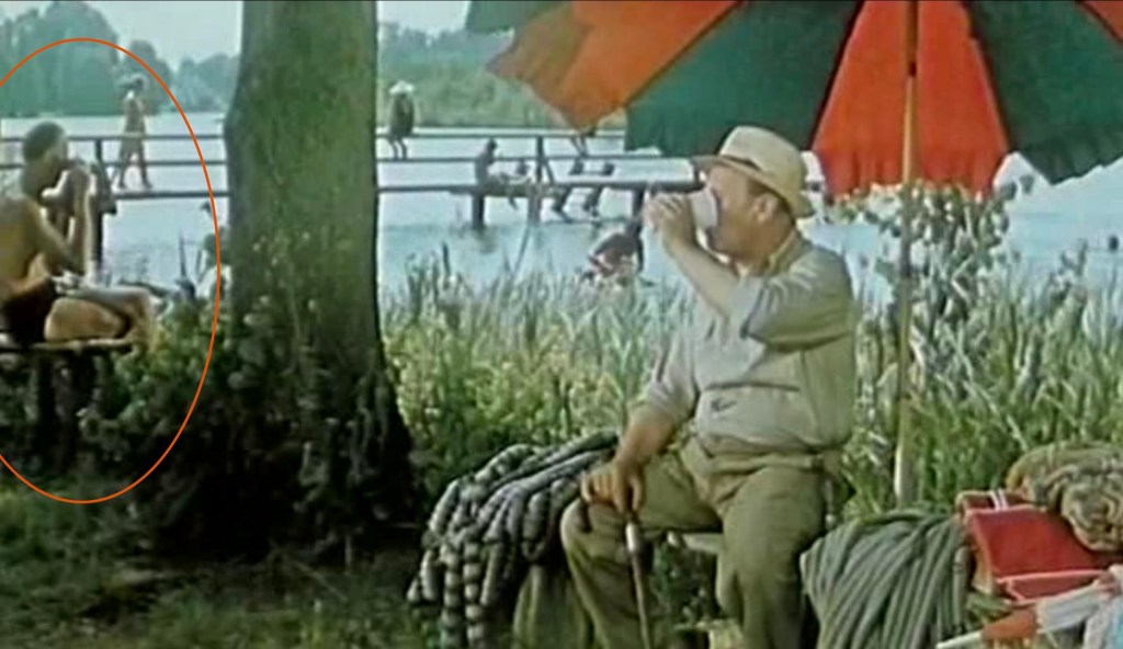 A man in the right foreground under a beach umbrella, with people bathing in a pond in the background. To the left, screenwriter František Hrubín is taking pictures.