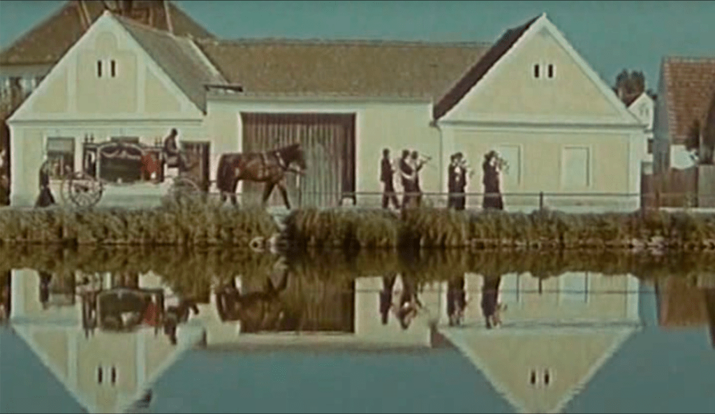 A procession of musicians and a horse carriage walks alongside a pond in front of a building. The scene is reflected in the pond.